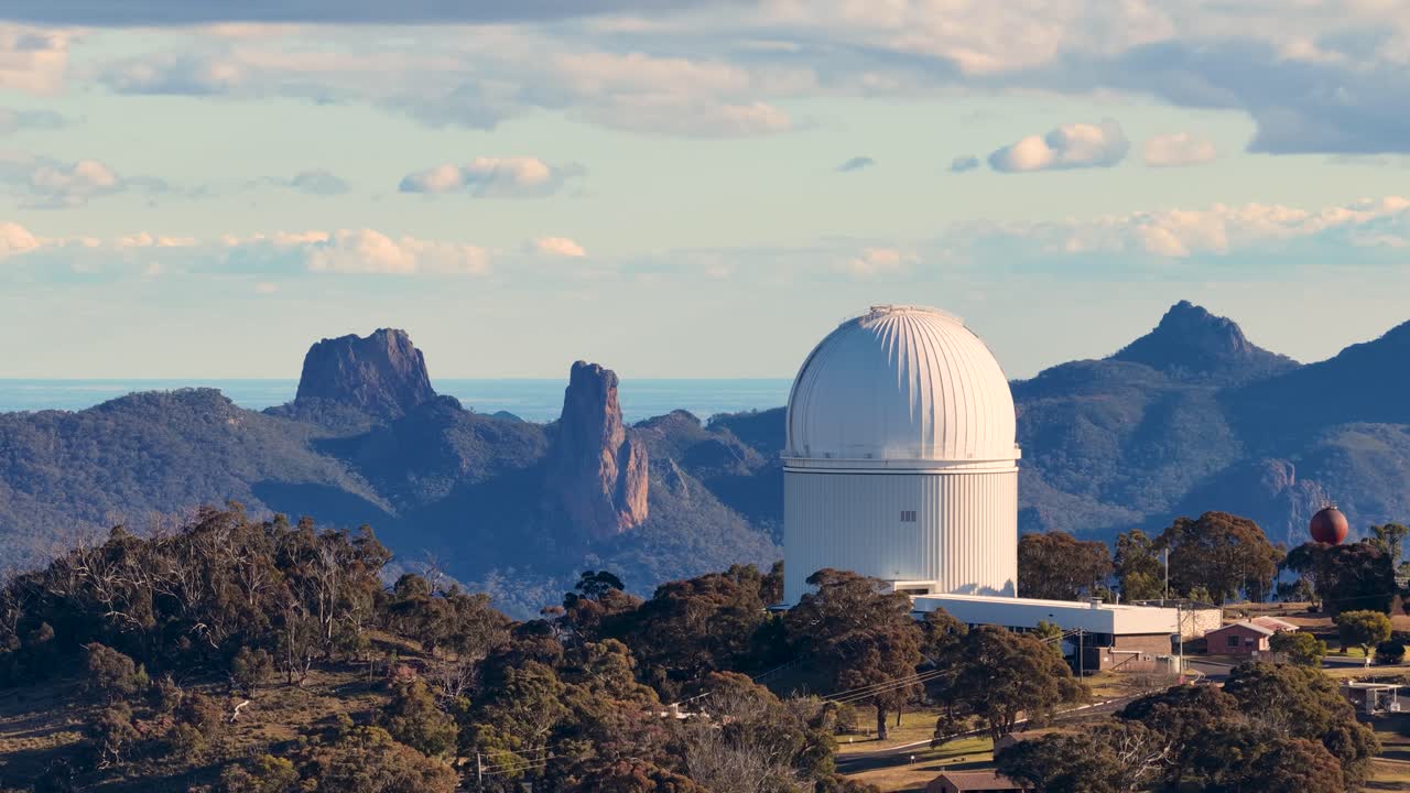 Drone camera slowly pans across a white observatory dome set among forested hills and rugged peaks under soft daylight in Warrumbungle National Park