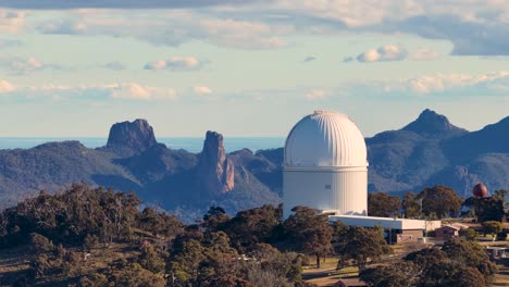 drone camera slowly pans across a white observatory dome set among forested hills and rugged peaks under soft daylight in warrumbungle national park