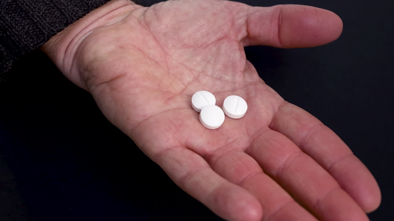 Video ravvicinato di una mano che si apre lentamente per rivelare tre pillole bianche appoggiate sul palmo. Concetto di farmaci, assistenza sanitaria, trattamento o uso farmaceutico, con uno sfondo scuro neutro