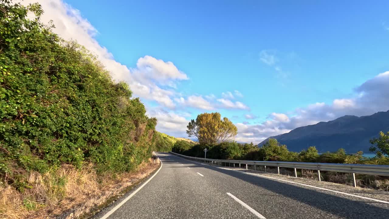 Car travels winding lakeside road, lush greenery, mountains, and blue sky in golden afternoon light