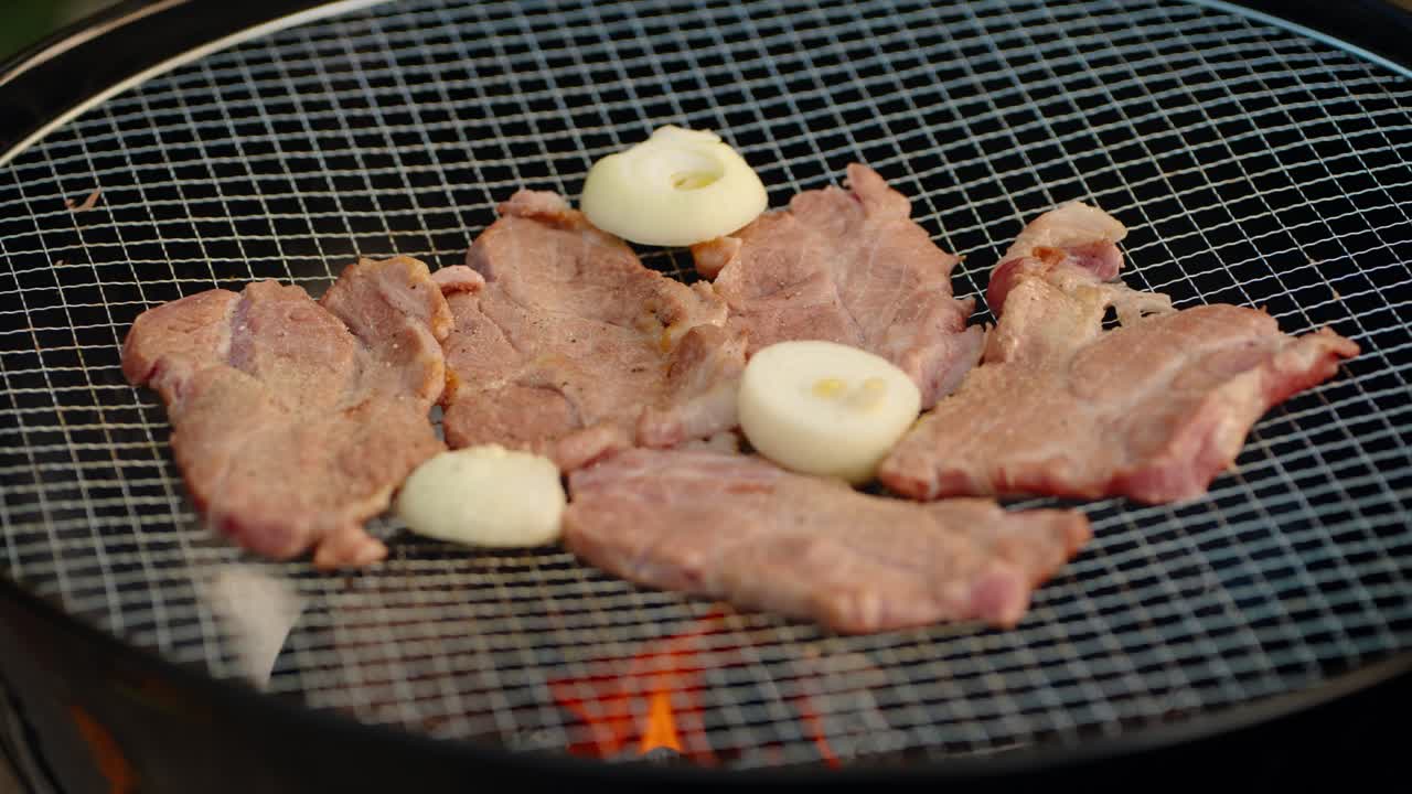 Thin slices of fresh pork and onion halves sizzle on a wire mesh grill over hot charcoal flames during an outdoor Korean barbecue, camping trip, or summer party