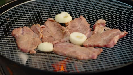 thin slices of fresh pork and onion halves sizzle on a wire mesh grill over hot charcoal flames during an outdoor korean barbecue, camping trip, or summer party