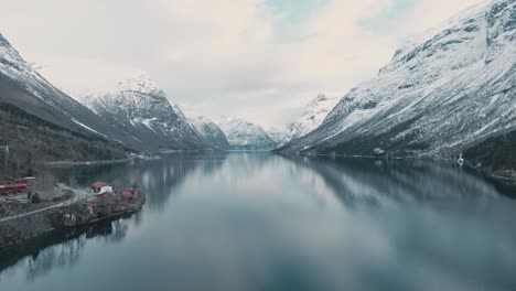 snow covered mountain peaks and crystal clear lake with small town in norway, aerial ascend view