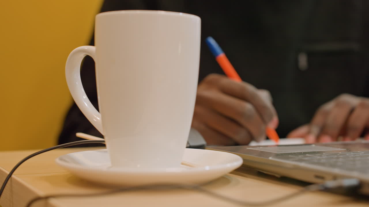 Mani di colore che scrivono vicino a un laptop con tazza di caffè in primo piano, movimento della penna arancione-blu, atmosfera accogliente da caffè, freelancer che fa brainstorming durante la pausa mattutina, toni caldi e messa a fuoco ridotta catturano un momento casuale