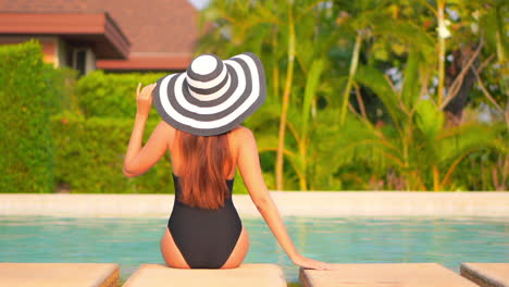 woman sitting on the edge of the swimming pool at an exotic hotel in florida wearing black monokini and striped hat and touching hat border, back view slow-motion handheld