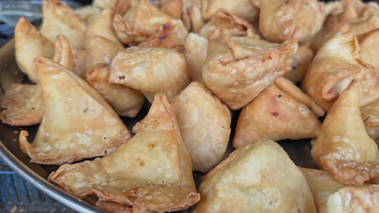 Carrellata su samosa appena fritte ammassate, pasta dorata e croccante che mostra bolle e pieghe, toni caldi che evidenziano la texture e l'abbondanza su un vassoio di metallo. dall'alto