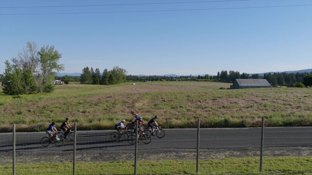 Gruppo di ciclisti che pedalano lungo una strada rurale delimitata da campi aperti e un fienile in lontananza.