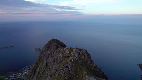 Hikers-standing-on-the-scenic-mountain-of-Reinebringen-in-Lofoten-enjoying-midnight-sun-during-summer,-Aerial-Orbit-shot