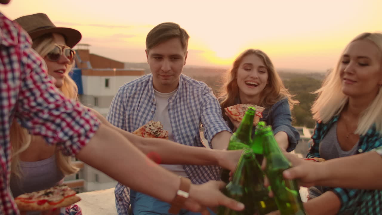 Young people clinks glasses and drinks beer from green bottels on the party with friends on the roof at the sunset.They eat hot pizza after in summer everning.