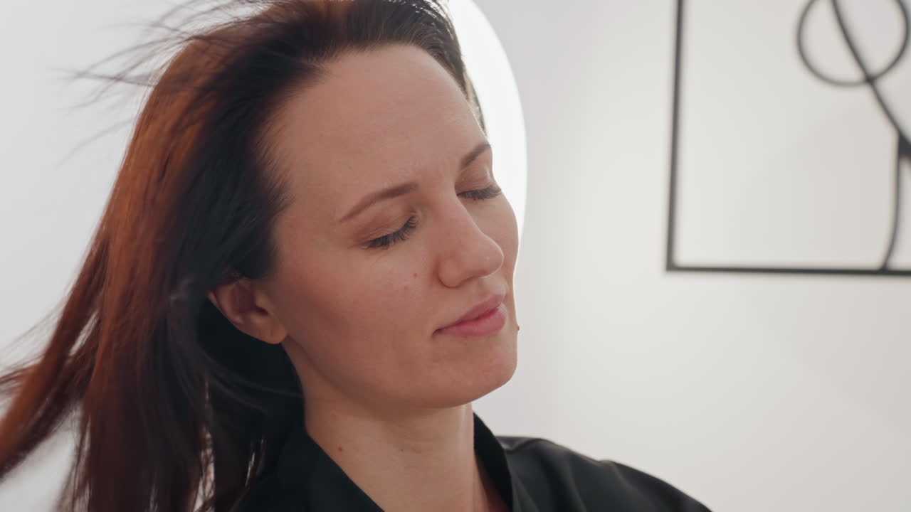 Donna con espressione riflessiva, stilista professionista che valuta il colore e la lunghezza dei capelli in studio, primo piano di una donna calma con capelli tirati indietro sotto la ring light per la valutazione del ritratto