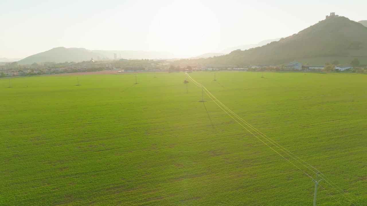Peaceful aerial view of vast green fields with power lines stretching into the distance as the sun rises over the horizon, highlighting the landscape's serene beauty and infrastructure. (4K)