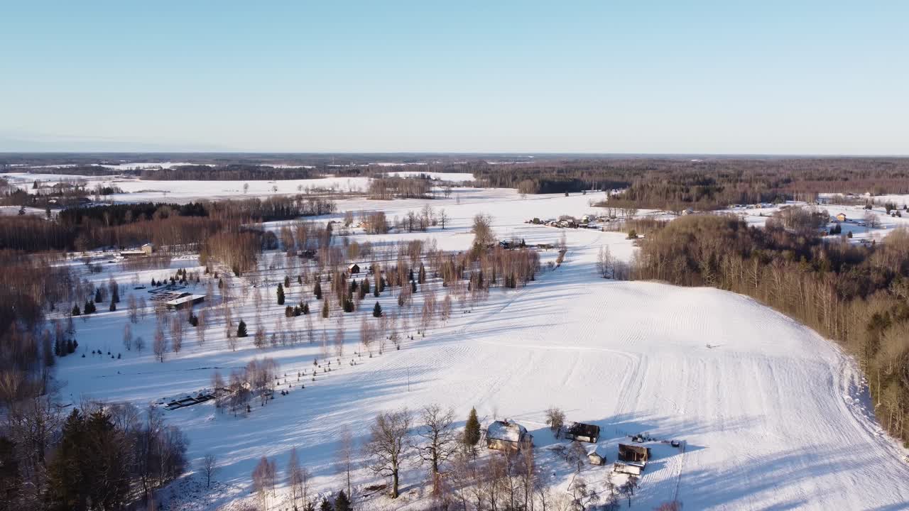 Aerial view of Krimulda, Latvia in winter showing snow-blanketed fields, rural homes, tree lines, and forest. Bright clear skies and open white terrain highlight serene seasonal stillness.