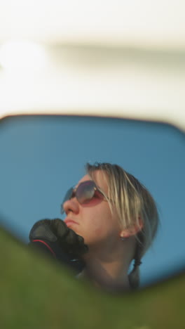 un reflejo de espejo de primer plano muestra a una mujer enfocada con gafas de sol, tocando suavemente su cara mientras mira hacia la distancia, el fondo revela un campo borroso y un cuerpo de agua