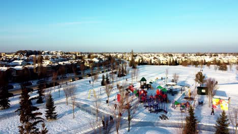 aerial j-flight backwards winter fly over snow covered childrens playground next to double loop skating track connected to an ice rink at birds eye view by homes at glastonbury park bon fire pits 2-2