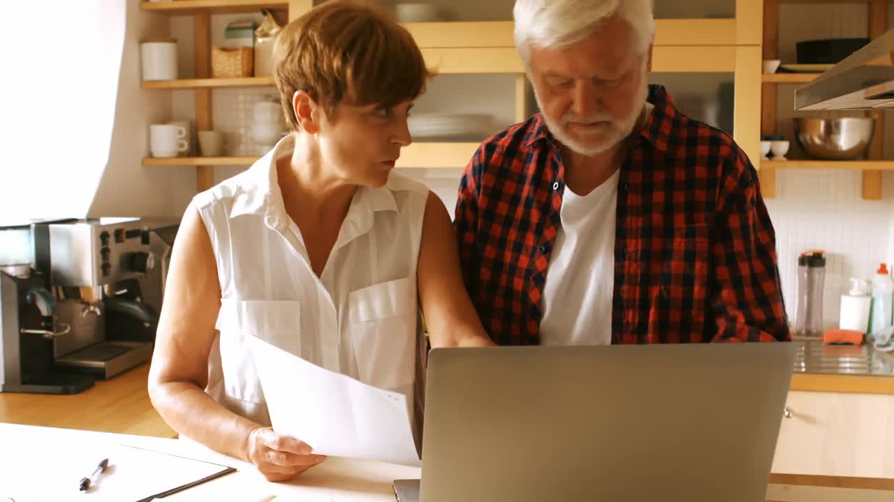 Senior couple paying bills online on laptop in kitchen