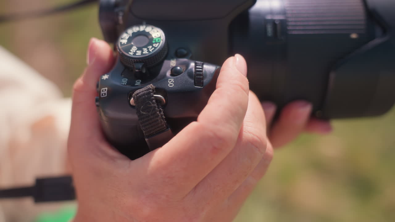 Close up view of fair skin hand adjusting DSLR camera settings with dial in natural light outdoors, lens in focus with soft blurred green background