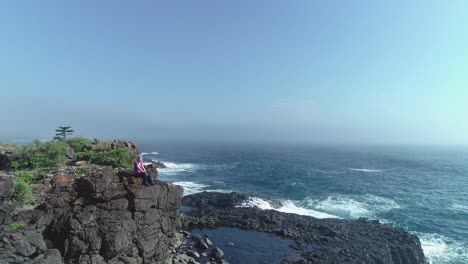 Aerial-drone-flying-forward-over-waves-breaking-on-rocks-pass-woman-sitting-on-cliff-towards-Kiama-Rock-Pool-and-out-to-sea-on-a-sunny-misty-morning