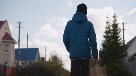 back view of person in blue jacket and black beanie strolling in residential area holding brown paper bag, surrounded by colorful houses, trees, and bright cloudy skies on a tranquil day