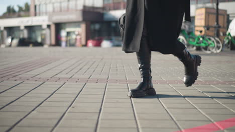 side leg view of individual in black coat and net trouser, walking briskly across an interlocked path, with a parked green bicycle and urban buildings blurred in the background