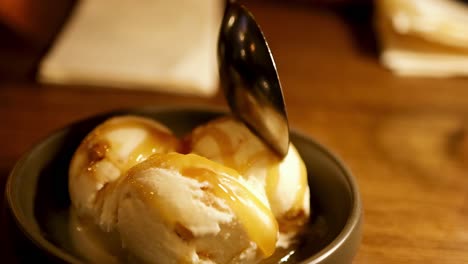 close-up of a spoon scooping creamy ice cream topped with caramel sauce from a bowl on a wooden table.