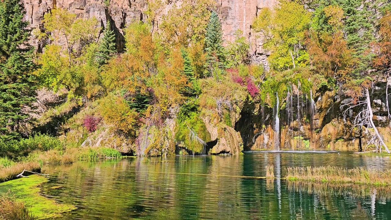 Vista panorâmica próxima das cachoeiras em Hanging Lake, Colorado, fluindo de musgo verde espesso e saliências rochosas para águas azul-turquesa, emolduradas por folhagem de outono e vegetação de cânion iluminada pelo sol