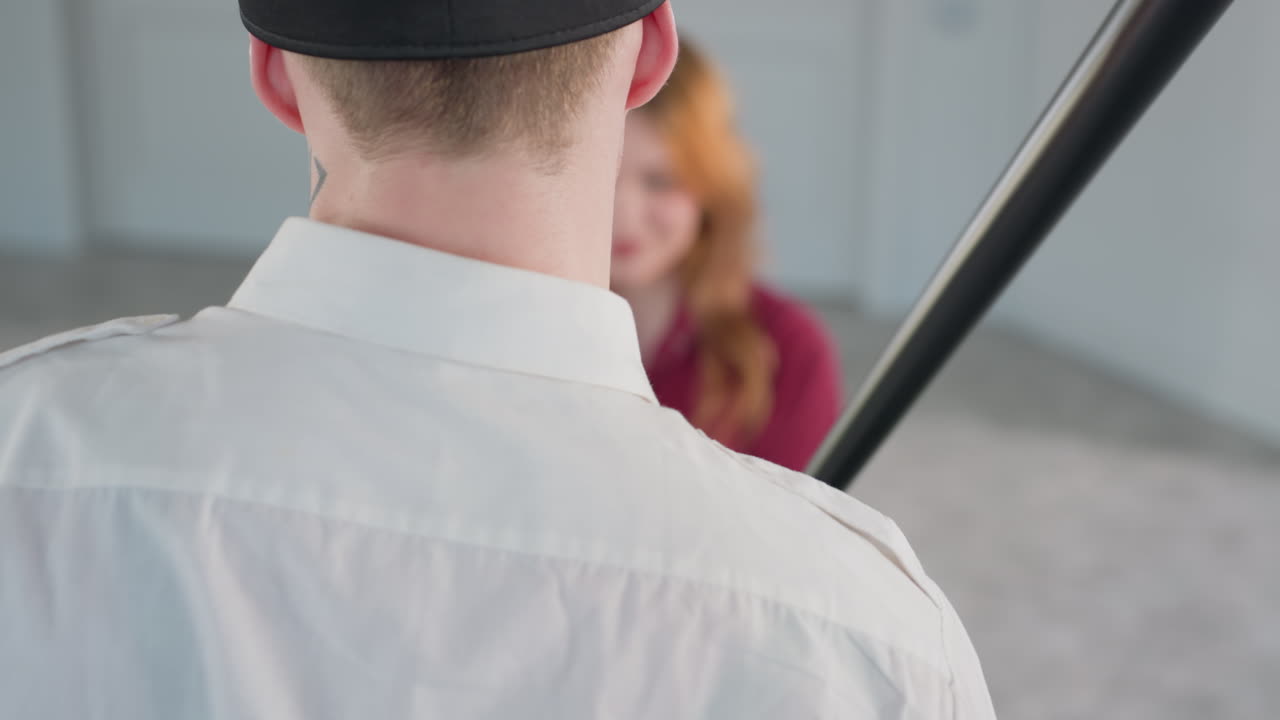 Rear view of man in white shirt holding black stick, playfully pointing it toward seated woman in red shirt, her face blurred in background, inside brightly lit modern interior with white walls