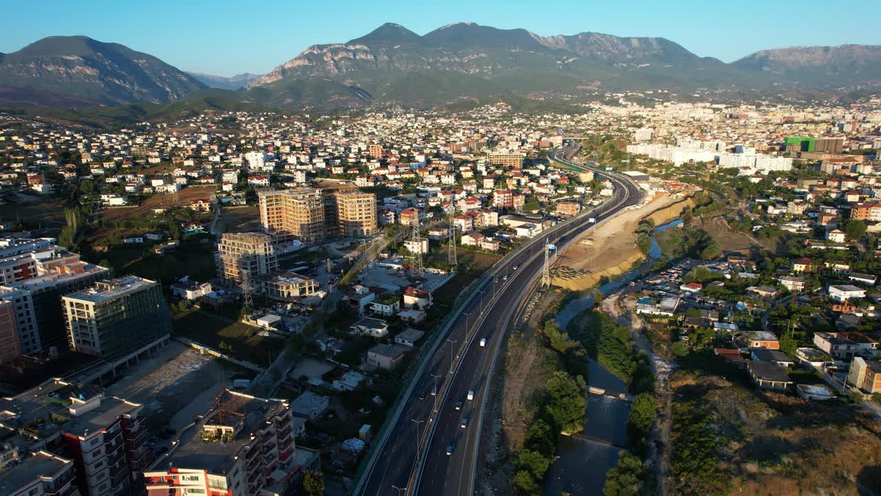Suburban Tirana at twilight with buildings, mountains in background, moving cars and people on infrastructure roads