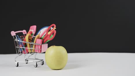 close up of shopping trolley with school items and apple with copy space on black background