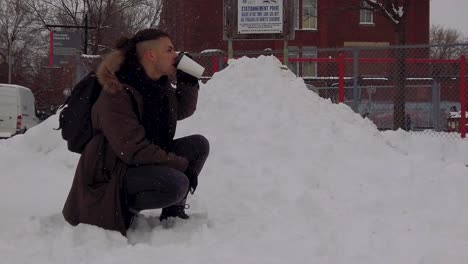 young man kneeling in snow taking drink of hot coffee