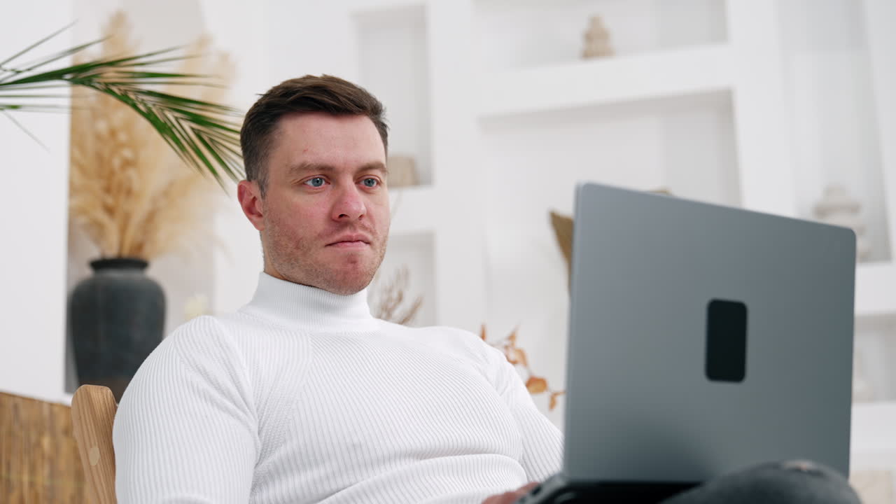 Serious Caucasian man wearing white shirt looks intently on his laptop. Comfortable workplace remote from the office. Low angle view.