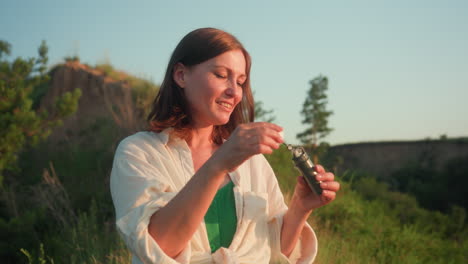sunset lit young woman in green skirt standing on grassy river bank blowing shimmering soap bubbles while smiling and gazing at floating spheres against blurred valley and flowers