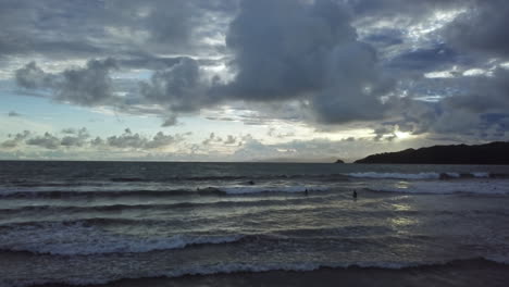 Vista-Aérea-Moviéndose-Hacia-El-Mar-Y-Nadadores-Al-Atardecer