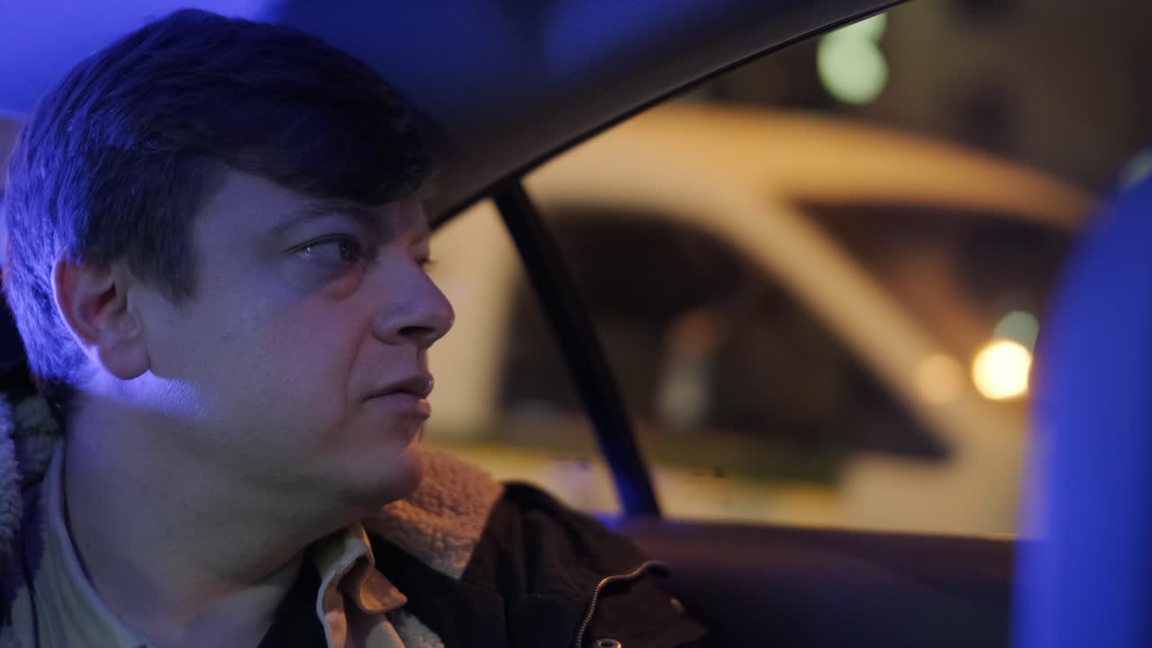 멋진 젊은 사업가의 느린 동작 클로즈업은 밤에 도시의 중심에서 운전자와 함께 현대적인 차로 여행하는 동안 밖을 바라보고 있습니다.