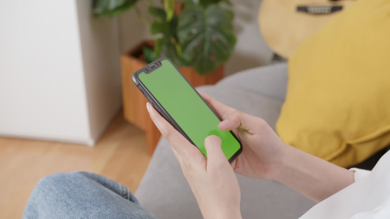 Donna che tiene in mano un mockup di smartphone con schermo verde sul divano di casa