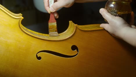 skilled luthier varnishing wooden cello center bout and ribs with traditional brush, highlighting meticulous handcrafting technique in professional instrument workshop, cremona, italy