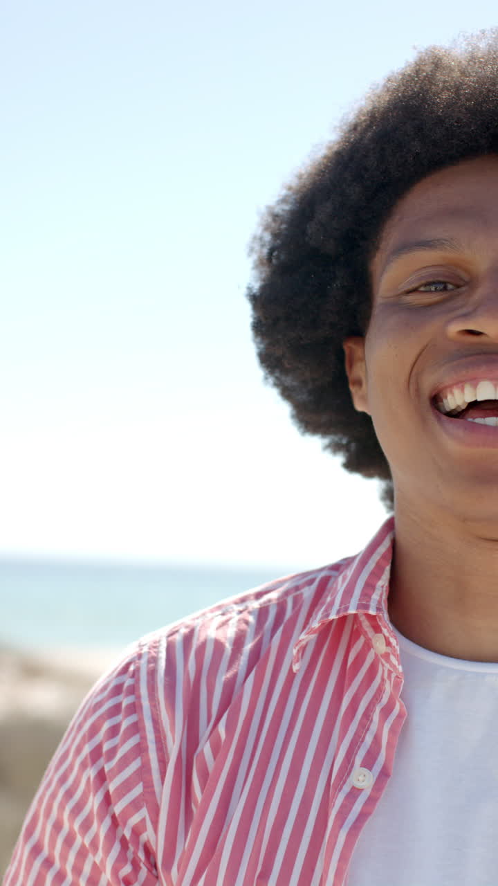 Vertical video: Smiling African American man enjoying sunny day at beach, wearing striped shirt