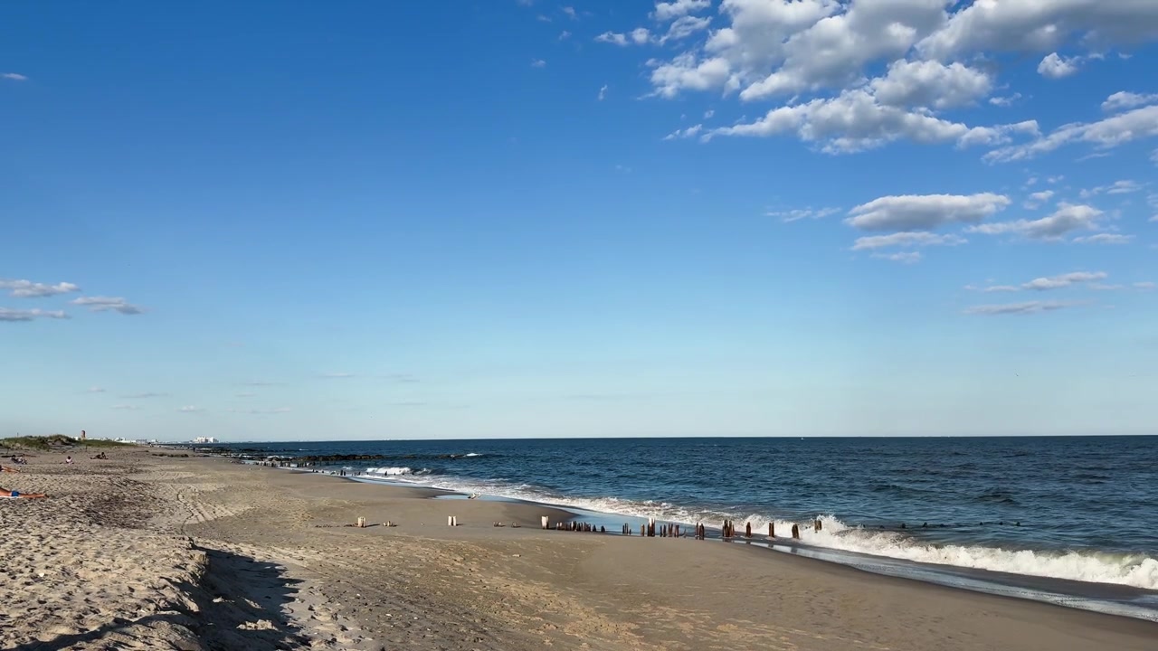 Ombak dengan lembut membasahi pantai berpasir yang luas di bawah langit biru cerah dengan awan.