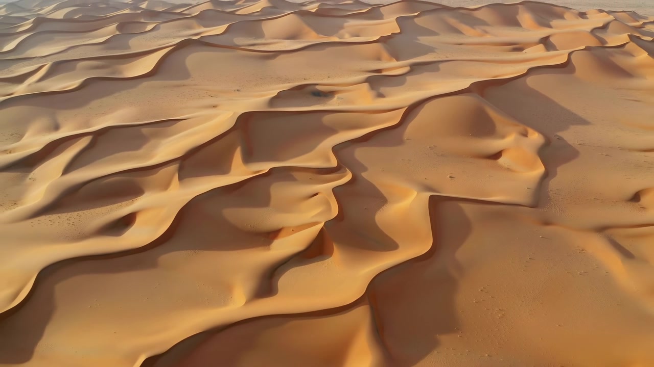 Vista aerea di morbide dune di sabbia desertiche che riempiono l'inquadratura, con creste stratificate, curve morbide e una calda luce solare che crea lunghe ombre attraverso il paesaggio aperto.