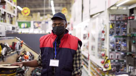 Salesman-in-mask-standing-between-rows-in-hardware-store,-looking-to-the-camera