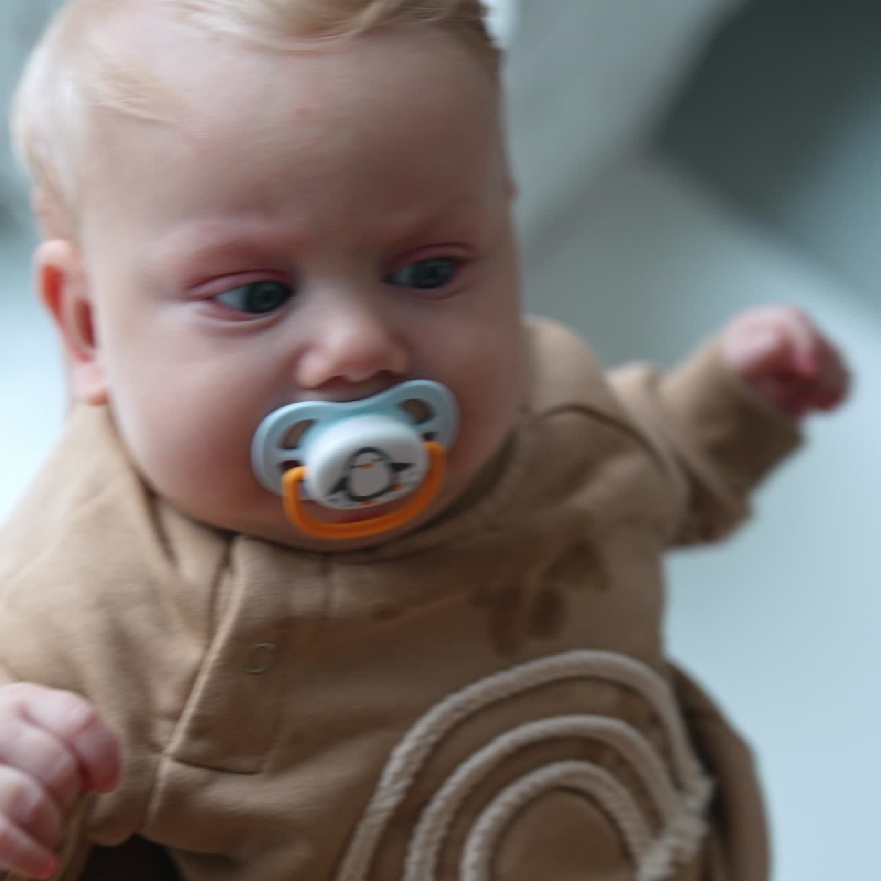 Söt blond liten pojke med stora blå ögon vinkas i händerna. Barnet suger på en napp och tittar uppmärksamt på mamma.