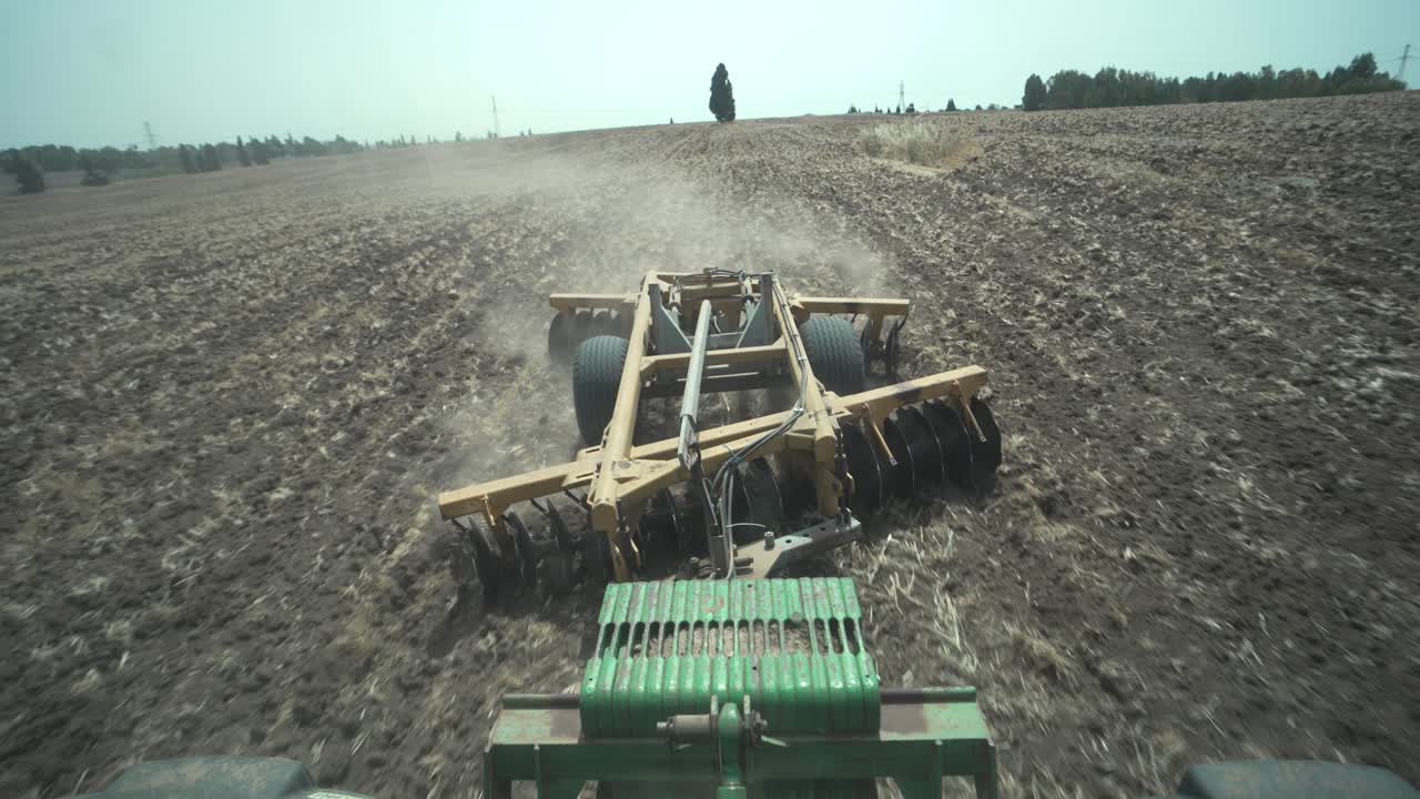 A disc harrow plows a dry field under bright daylight, raising dust clouds. Blue summer skies. Field ready for sowing. Shot from tractor driver POV. Modern agriculture in Israel, the Holy land