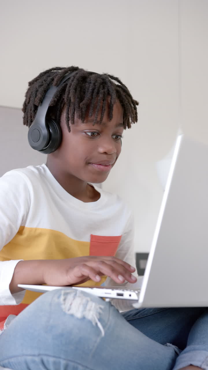 Vertical video of happy african american boy sitting on bed using laptop at home, slow motion