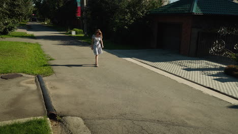 fairs skinned lady in floral dress takes relaxed stroll along peaceful neighborhood street during sunny day, green lawns, trees, and residential buildings line background