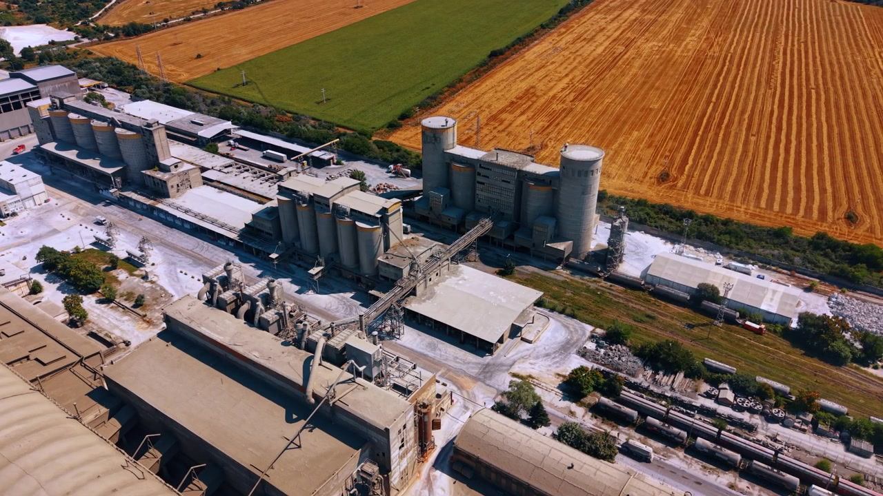 Zementlagersilos neben abgeernteten Feldern. Hochwinkel-Luftaufnahme von zylindrischen Türmen, Fabrikdächern, Förderbändern und Eisenbahnlinien nahe orangefarbenem Ackerland.