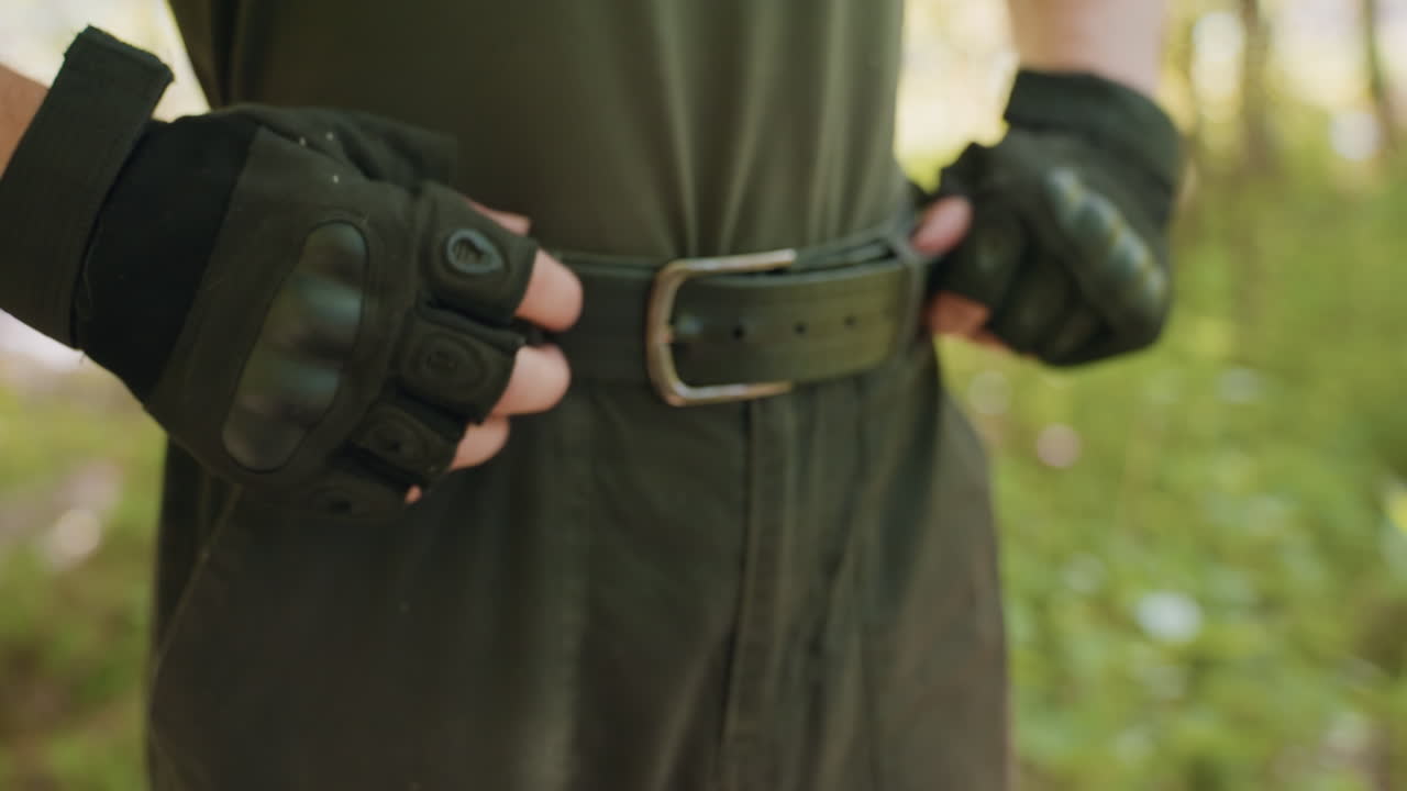 Waist shot of warrior in sleeveless top adjusting black leather belt and gloves amid sunlit forest, emphasizing tactical gear detail and rugged attire, under canopy of leafy woodland backdrop