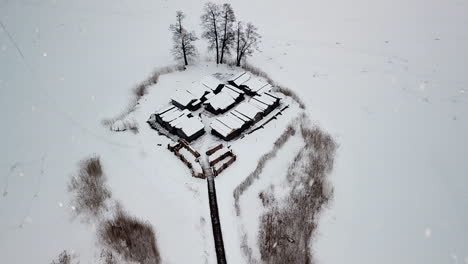 snow covered wooden buildings surrounded by white frozen lake while snowing, aerial drone view