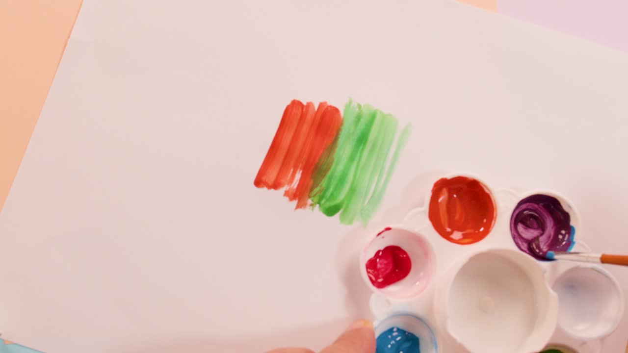 Paintbrush dips into purple paint beside colorful brushstrokes on white paper, overhead view