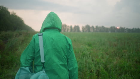 back view of individual in raincoat carrying bag and umbrella walking through misty green field, framed by scattered bushes and trees under an overcast sky with a distant sun peeking through clouds