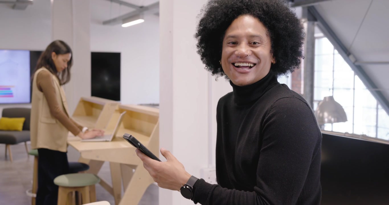 Afroamerikanske kolleger på åbent kontor, mand griner mens han læser på telefonen, kvinde taster på bærbar computer