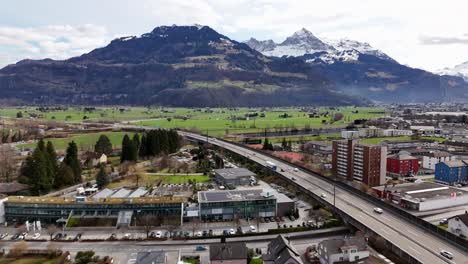 Viel-Befahrene-Autobahn-In-Idyllischer-Schweizer-Stadt-Und-Berge-Mit-Schneebedeckten-Gipfeln-Im-Hintergrund